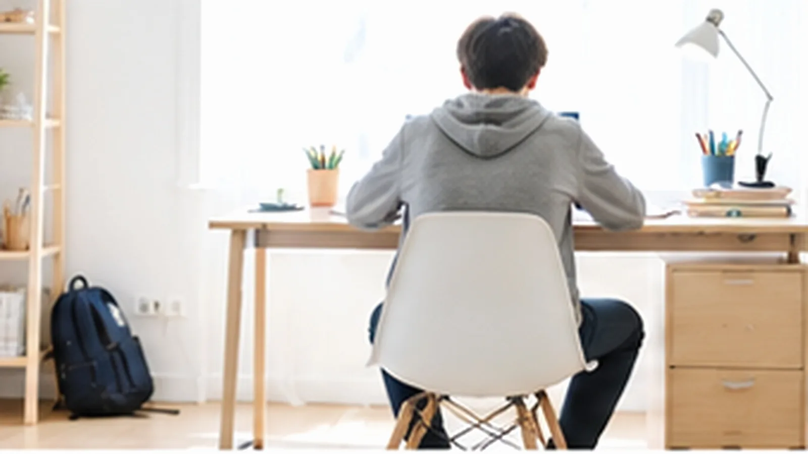 Un adolescent de dos, assis à son bureau dans une chambre claire aux murs blancs et au mobilier en bois